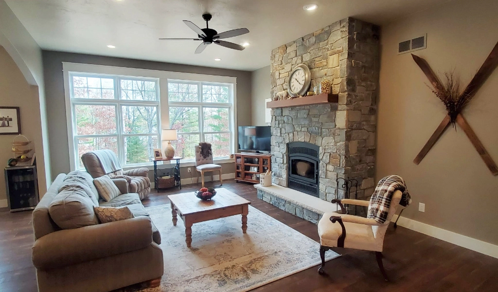 Living room with a large window, brown sofas, and a wooden coffee table. The stone fireplace features a clock, while the TV and armchair provide comfort. A ceiling fan spins overhead. For those considering home remodeling in Eagle River, the decorative wall piece adds character.