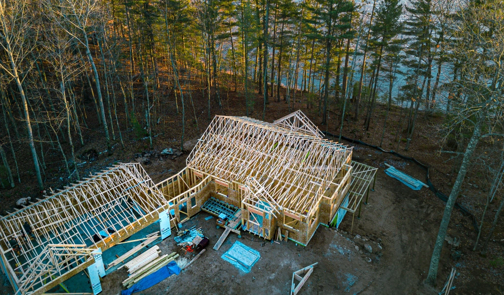 An aerial view reveals a house under construction in a forested area, showcasing the exposed wooden frame and roof trusses expertly crafted by a skilled design-build contractor in Eagle River, WI.