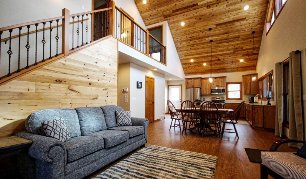 Spacious living area with wooden interior, featuring a gray couch, dining table with chairs, and a kitchen in the background. A staircase with a metal railing leads to the upper level, showcasing expert craftsmanship from a custom home builder.