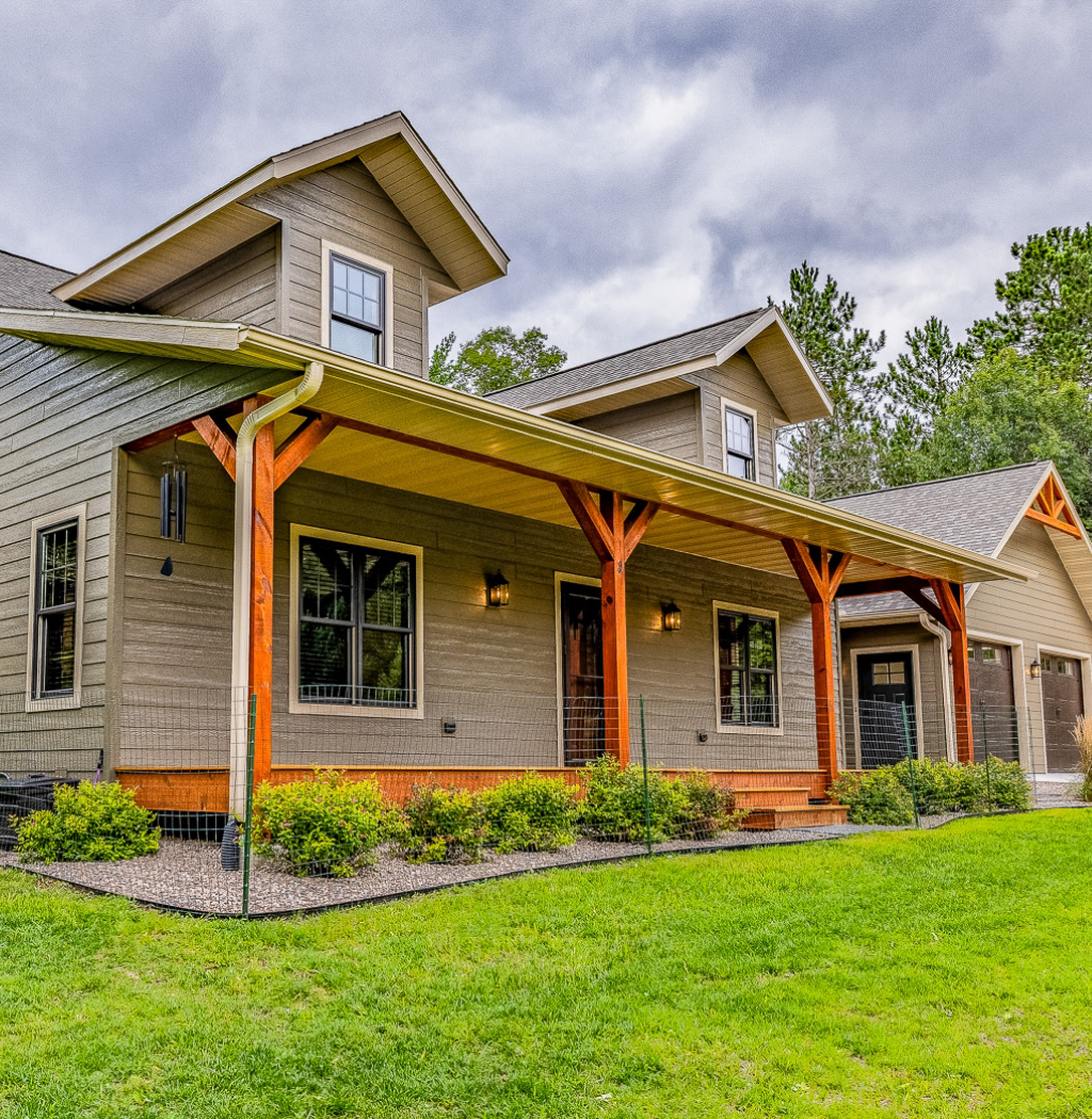 A modern house with a wooden porch, gabled roof, and two dormer windows, surrounded by a green lawn and trees, showcases the expertise of a design-build contractor. Perfect for those considering home remodeling in Eagle River.