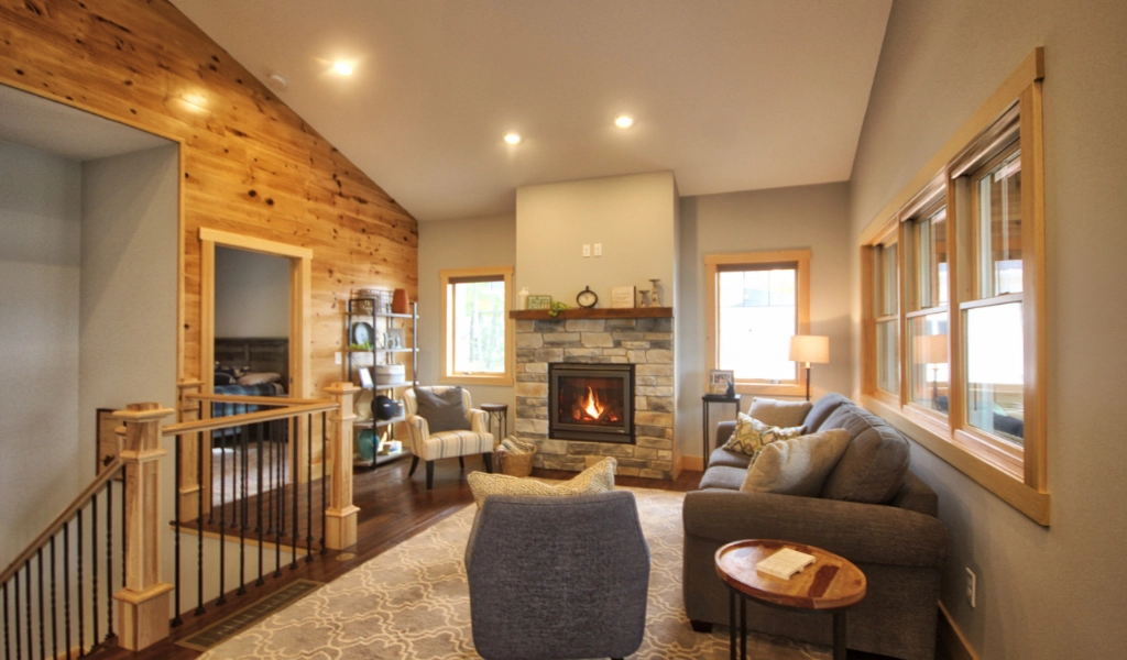 Cozy living room in this custom home construction features a stone fireplace, gray sofa, armchairs, and wooden accents. The pine-paneled wall and windows provide natural light, showcasing the craftsmanship of a design-build contractor near Eagle River, WI.