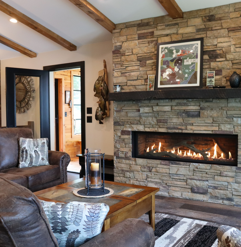 Nestled in the heart of a custom home construction project, this cozy living room features a stone fireplace, leather couches, and a wooden coffee table. A framed map graces the mantel, while a doorway opens to a wood-paneled hallway crafted by your trusted general contractor in Eagle River.