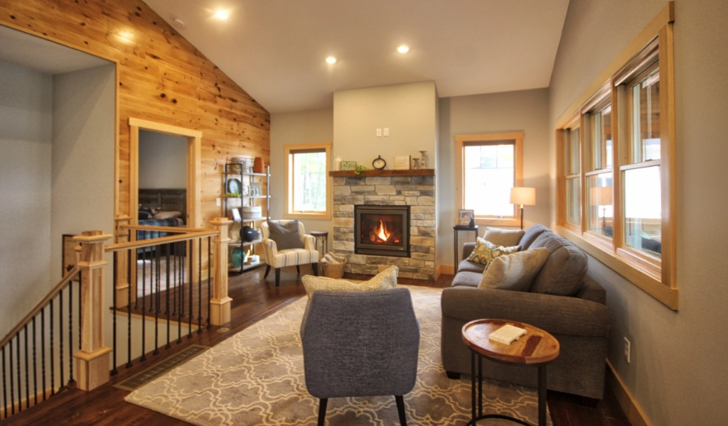 Cozy living room with a fireplace, gray sofa, two gray chairs, and a wooden coffee table. Large windows and recessed lighting brighten the space. Perfect for those considering home remodeling in Eagle River to create a warm, inviting environment.