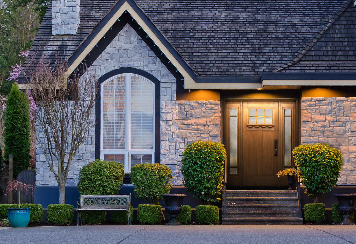 A house with stone siding, a gabled roof, a wooden door featuring glass panels, trimmed bushes, a small tree, and a bench in front.