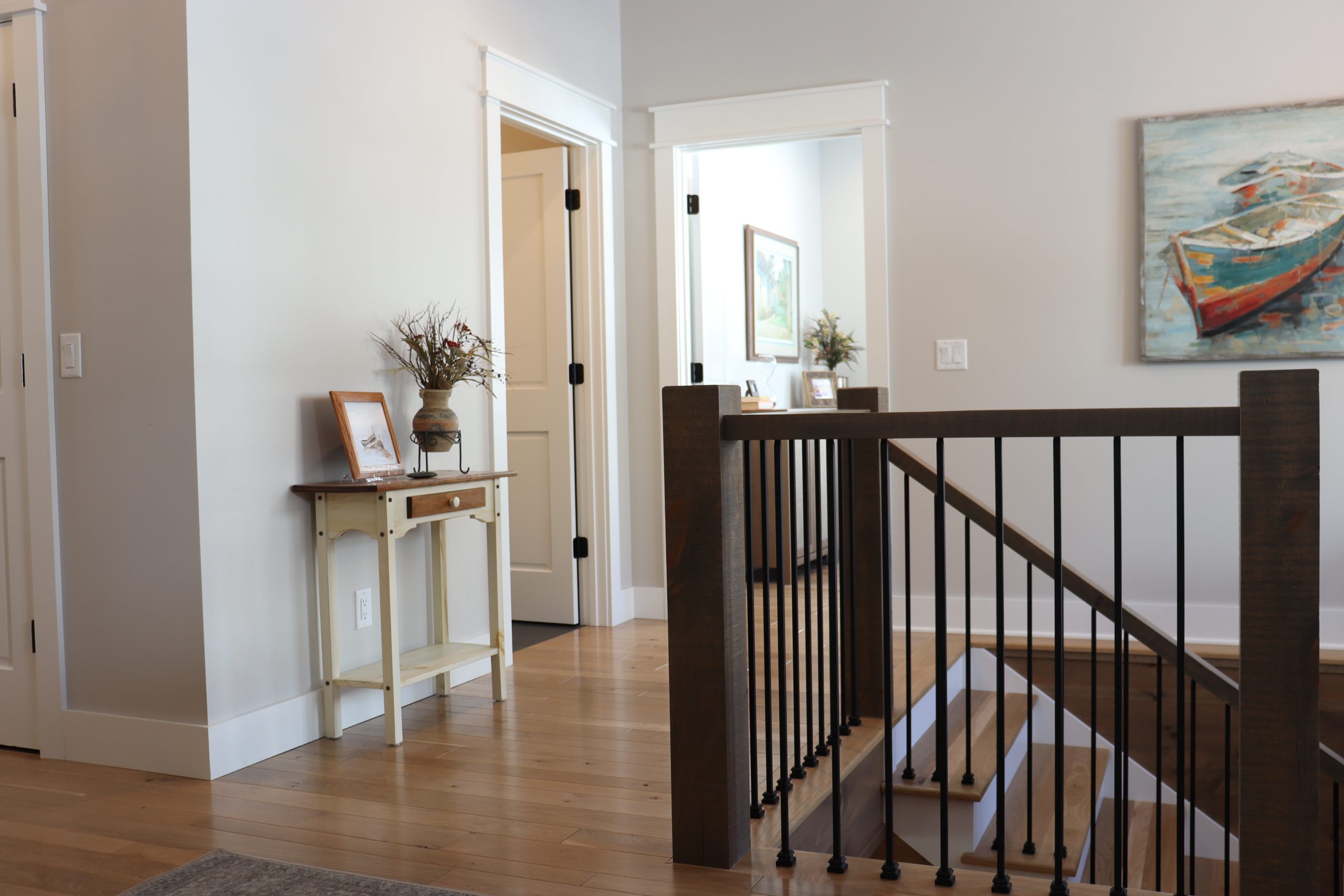 A hallway in a legacy home featuring light wooden flooring, a small table with decor items, white walls, and a staircase with dark railings.