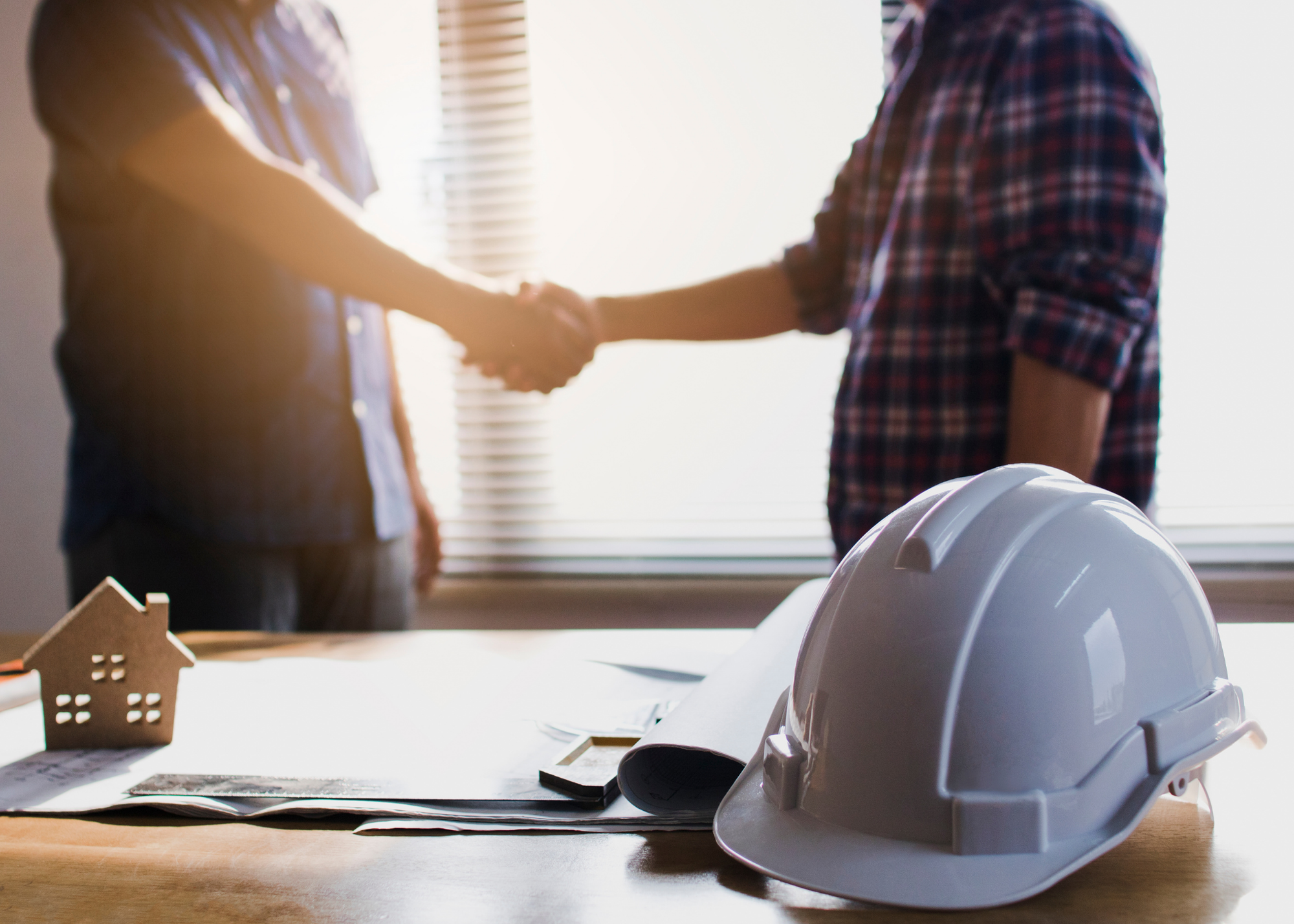 Two people shake hands indoors near a table with a white hard hat, blueprints, and a small house model, suggesting a construction or real estate meeting focused on design.