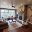 A living room with large windows framed by elegant trim, a stone fireplace, a beige sofa set, a wooden coffee table, and a wall-mounted TV. The decor features a rug, clock, and mounted skis for added charm.