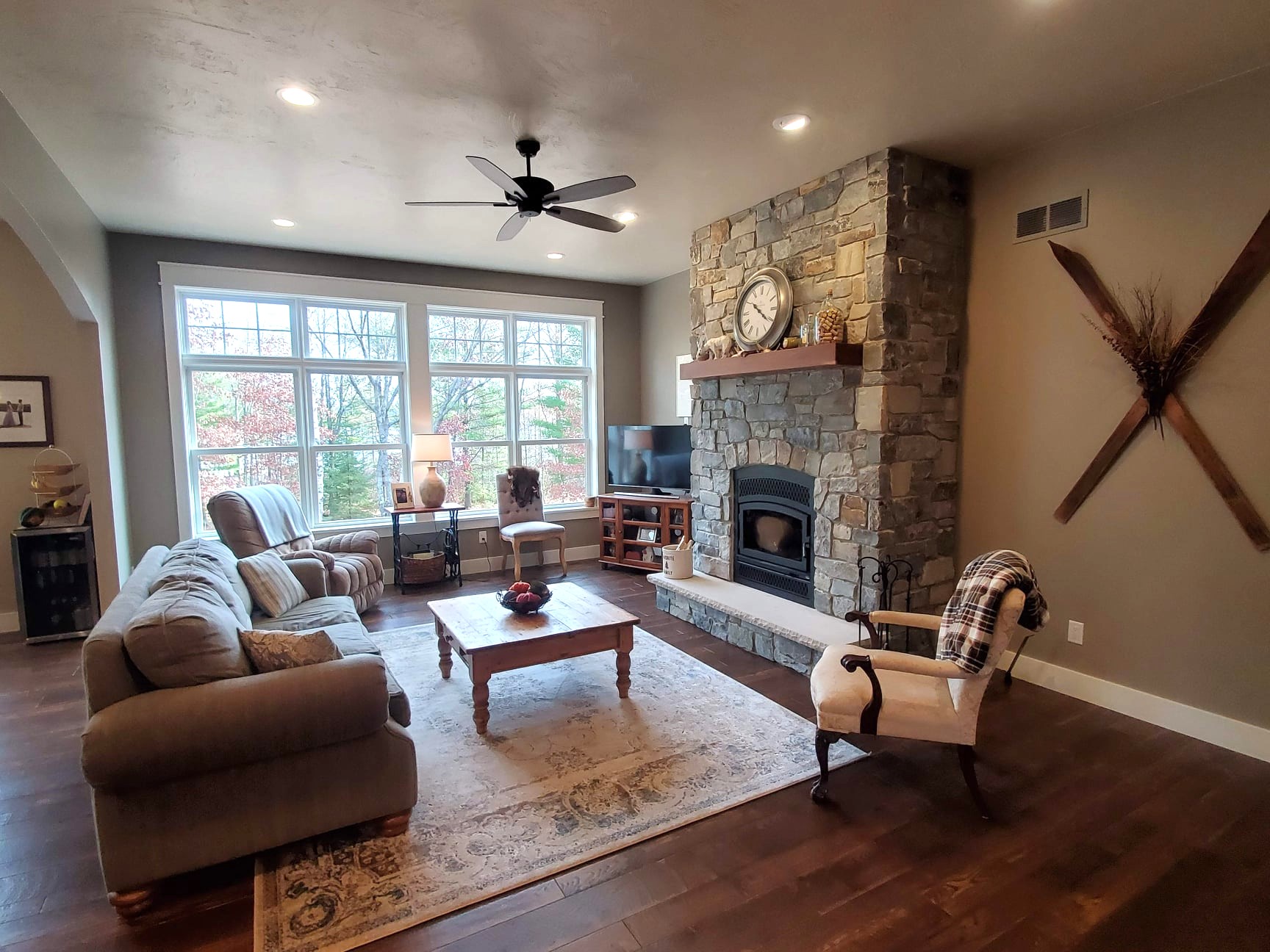 A living room with large windows framed by elegant trim, a stone fireplace, a beige sofa set, a wooden coffee table, and a wall-mounted TV. The decor features a rug, clock, and mounted skis for added charm.