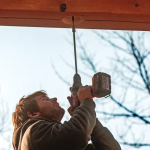A person uses a cordless power drill to drive a screw into a wooden beam outdoors, with tree branches in the background—a typical scene in Home Builder Jobs.
