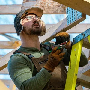 A man in work gloves and a hat stands on a ladder, using a pencil to mark a wooden beam at a construction site.