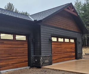 A modern black custom home with two wooden garage doors sits on a wet, unfinished driveway surrounded by trees and dirt, with a hint of stonework and a yellow container near the garage.