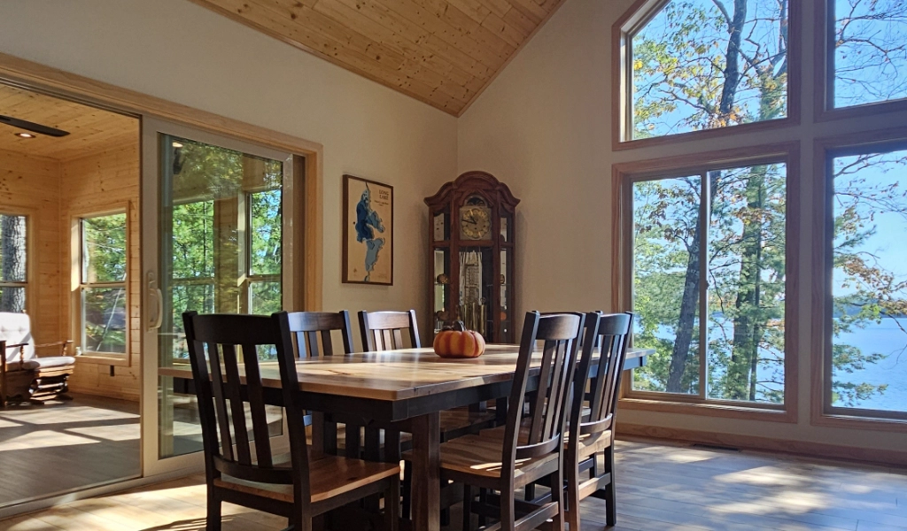 A wooden dining table with six chairs and a pumpkin centerpiece sits by large windows overlooking trees and a lake in sunlit Timbercrest Cove, featuring wood-paneled walls and ceiling.