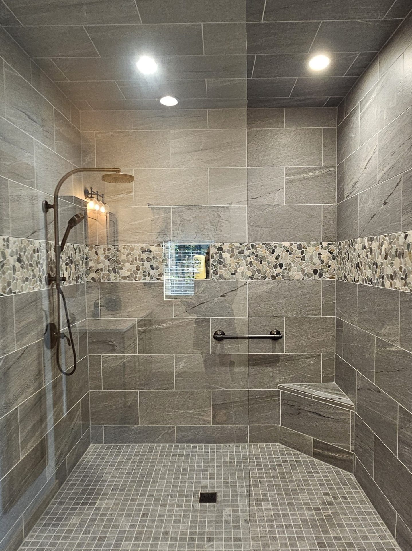 Modern walk-in shower with gray tile walls and floor, a mosaic accent strip, built-in bench, grab bars, glass door, and black shower fixtures—perfect for showcasing unique tile pattern selections in your new home.