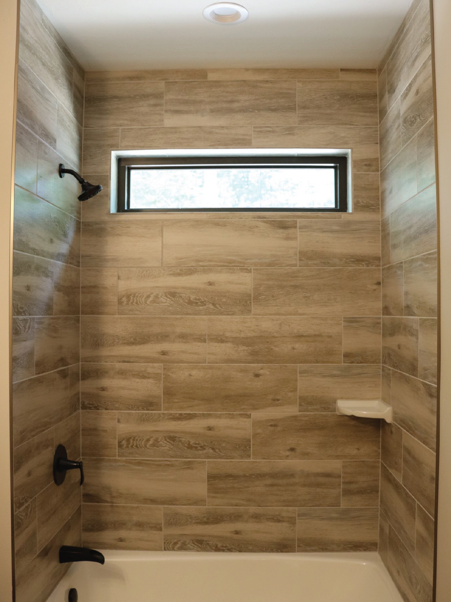 Shower with wood-look tile walls, a built-in shelf, matte black fixtures, a small rectangular window, and a white bathtub.