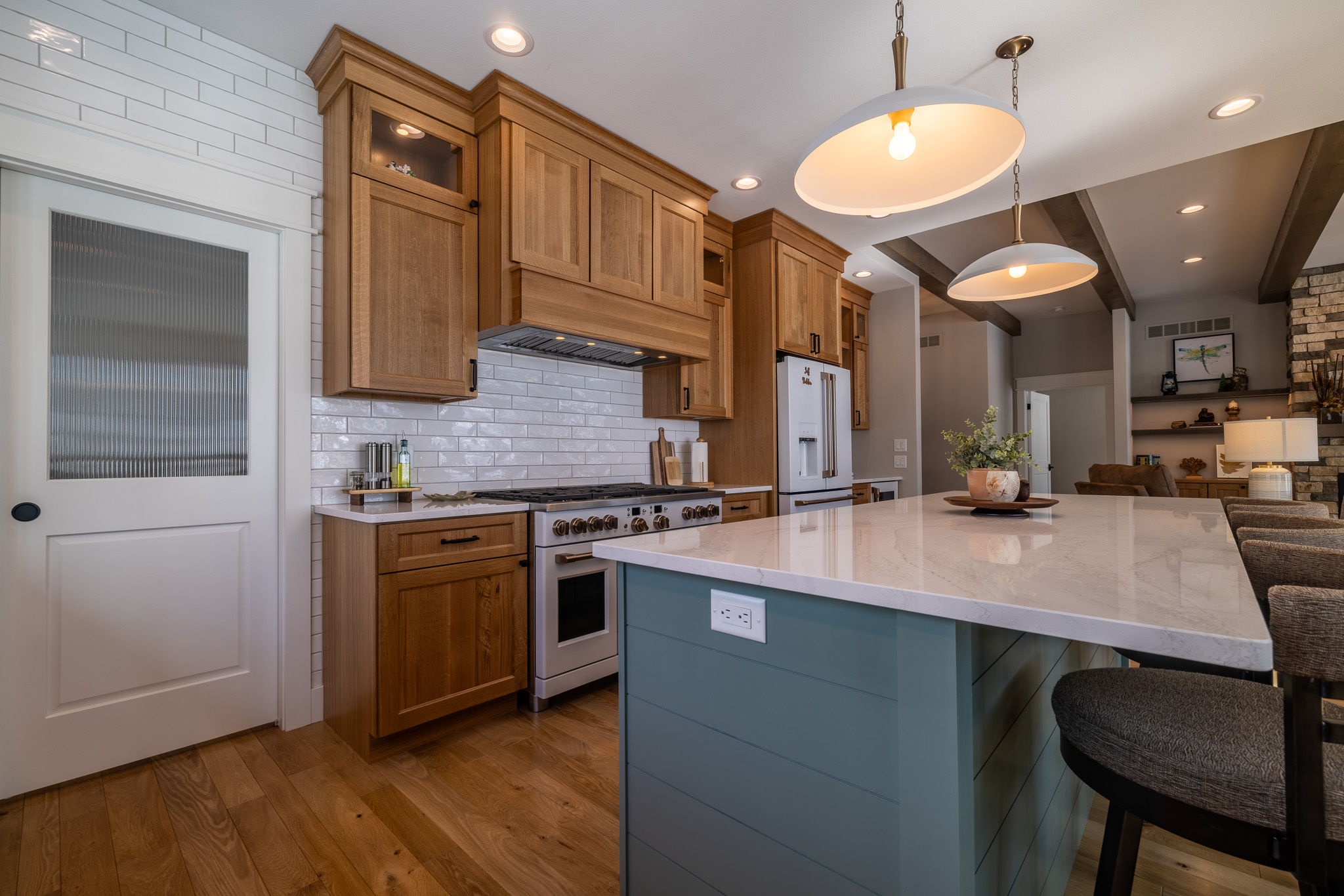 Modern kitchen with wood cabinets, white tile backsplash in a sleek tile pattern, island with marble countertop, pendant lights, and barstools; open layout with view into living area.