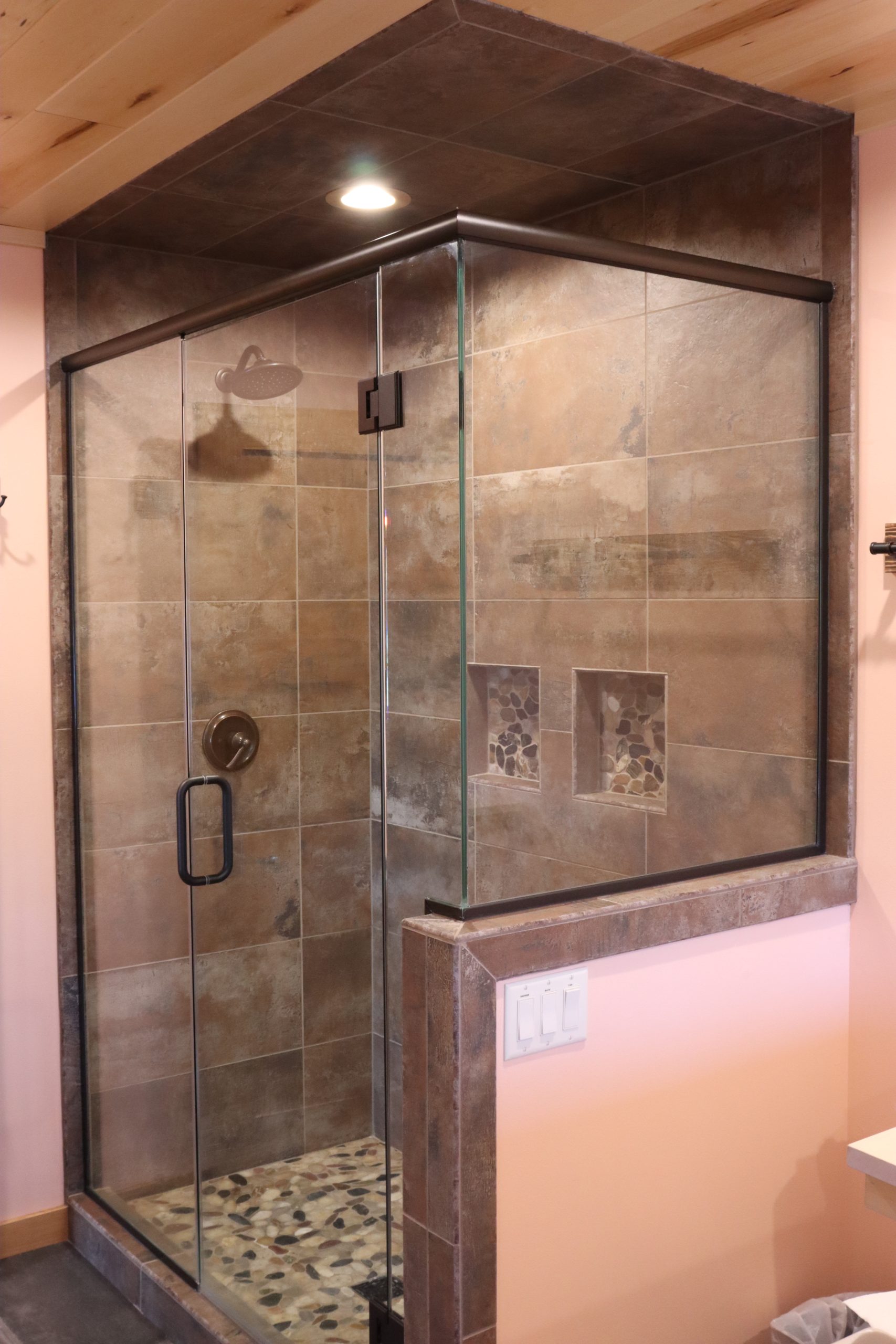 Glass-enclosed corner shower with a stylish tile pattern on brown tile walls, pebble stone floor, and built-in shelves, located in a bathroom with light-colored walls and wood ceiling.