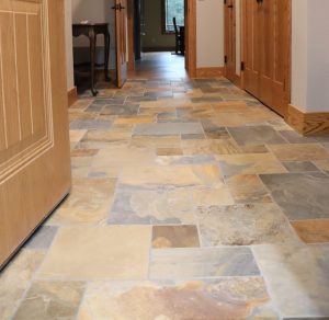 A hallway with a unique tile layout featuring multi-colored stone flooring, light gray walls, and wooden doors, leading to a room with a window and table—perfect inspiration for your new home.
