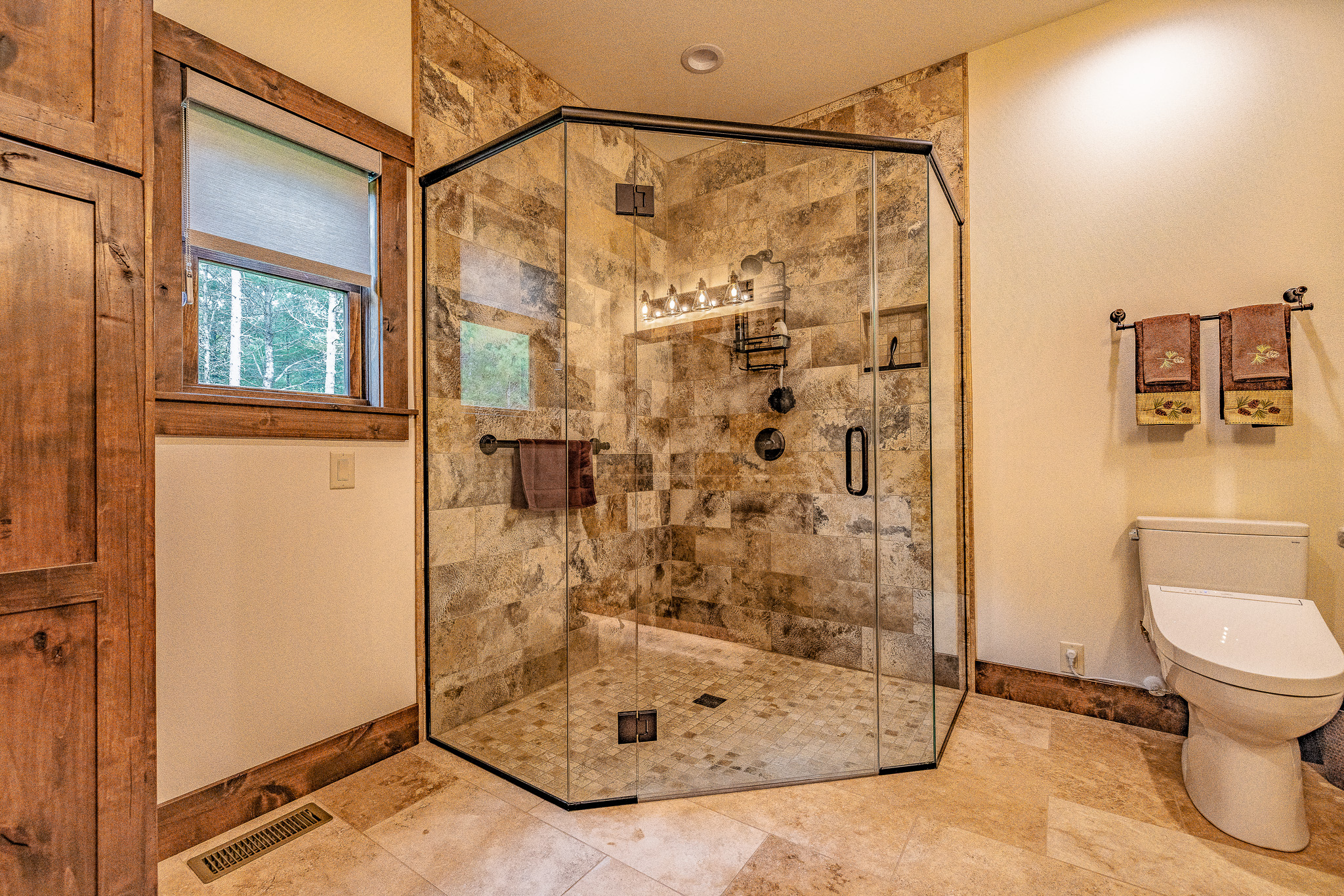 Spacious bathroom with a glass-enclosed walk-in shower, tiled walls and floor showcasing stunning tile pattern selections, a window, wooden accents, and a toilet with two towel racks above it—ideal for your new home.