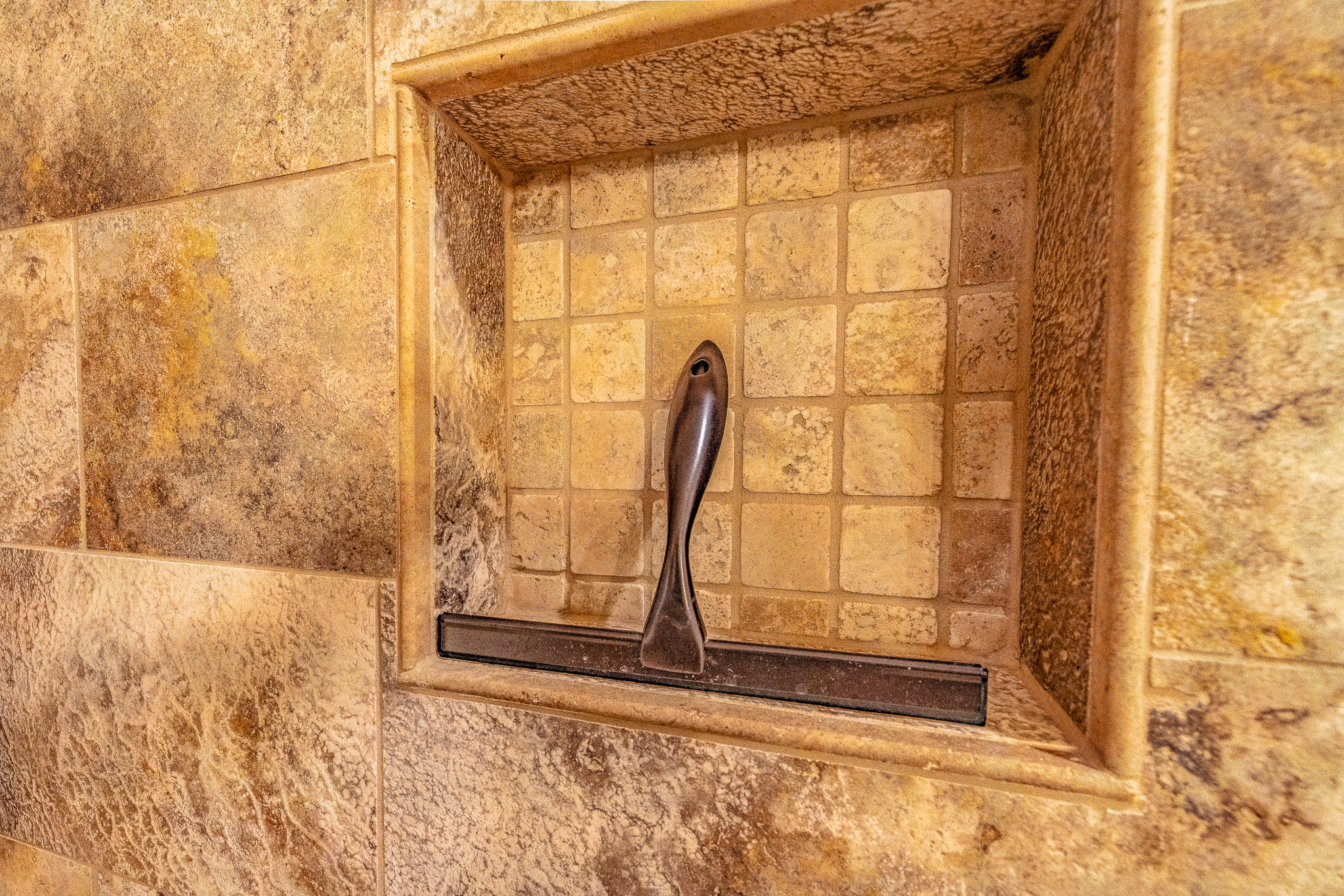 A metal squeegee is placed upright in a recessed tiled niche on a stone-tiled shower wall, showcasing thoughtful tile layout and stylish tile pattern selections in this new home.