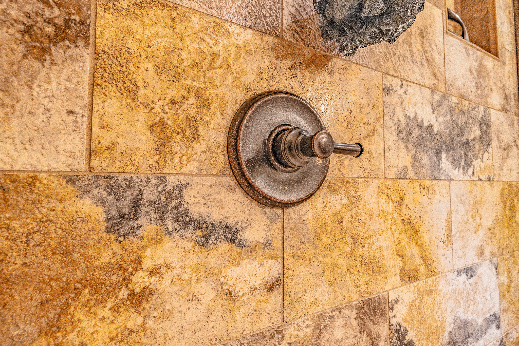 A metal shower handle is mounted on a wall of yellow, brown, and gray stone tiles, showcasing an elegant tile layout. A dark bath pouf hangs in the upper right corner—an inviting touch for any new home.