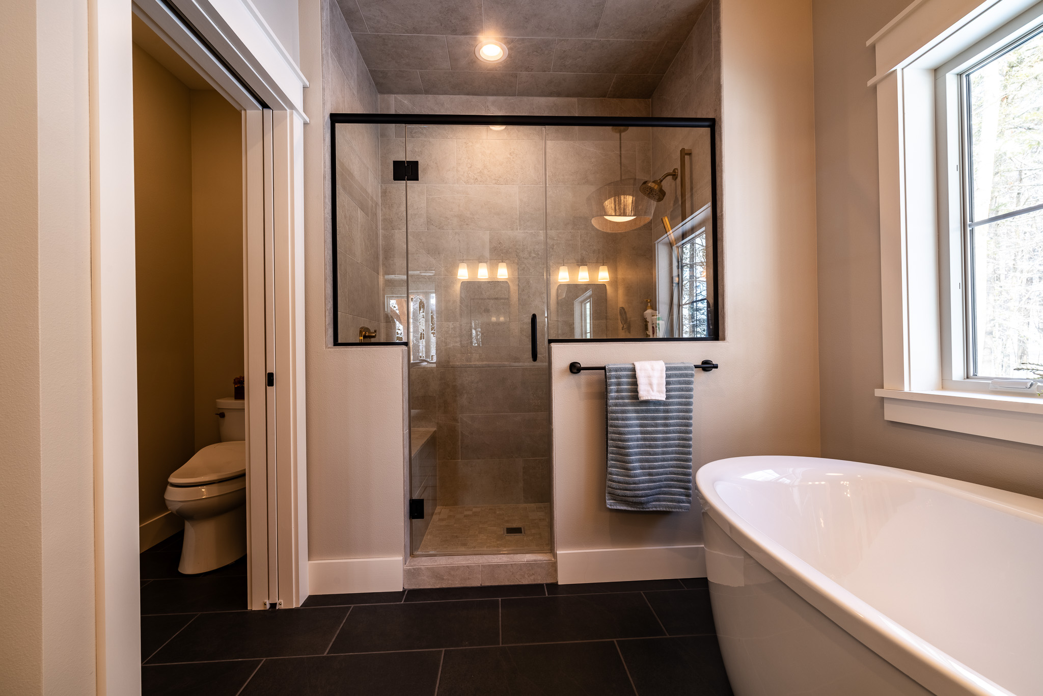 Modern bathroom with a glass-enclosed custom tile shower, freestanding bathtub, towel on a rack, and separate toilet area, featuring neutral tones and natural light from a window—perfect for anyone designing tile showers.