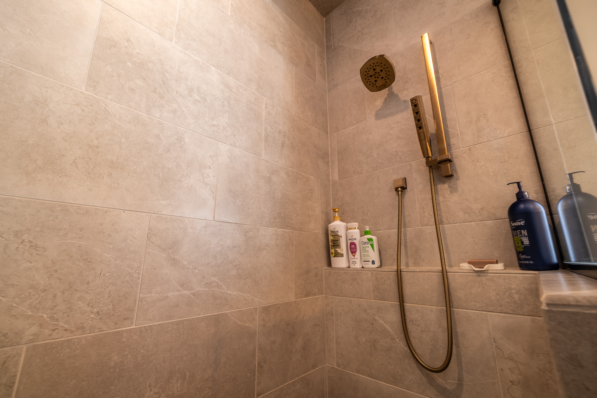 Modern shower with beige tiled walls showcases expert Tile Shower Design, featuring a gold showerhead and handheld sprayer, three bottles of toiletries, and a soap dish on a small ledge.