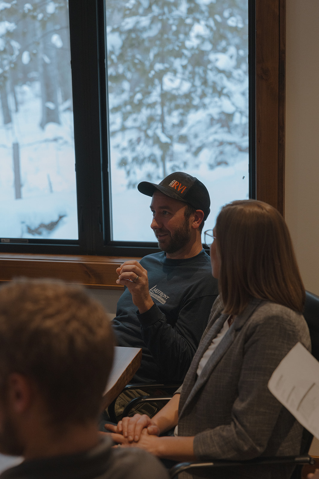 A man in a black cap speaks while sitting beside a woman during a meeting in a room with snowy landscape visible through the window, where custom tile showers are being discussed.