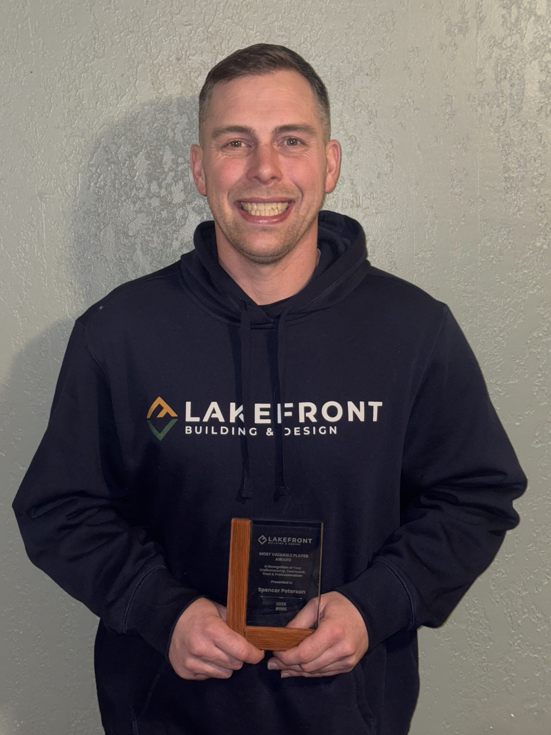 A man wearing a "Lakefront Building & Design" hoodie stands against a gray wall, smiling and holding a plaque award.