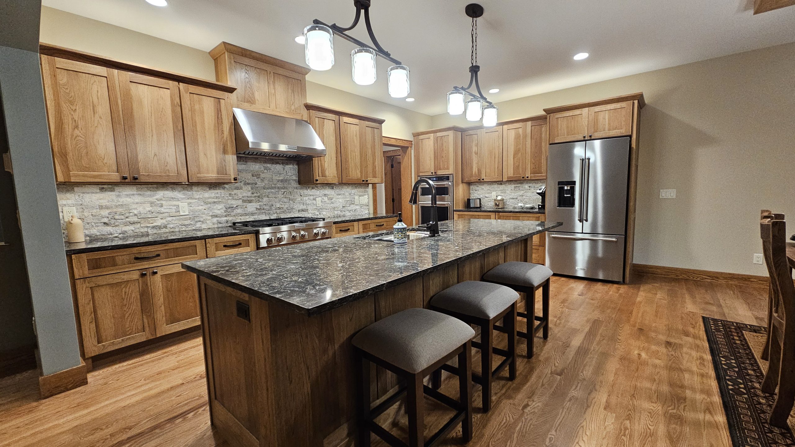Modern kitchen with wooden cabinets, stainless steel appliances, a large granite island with three stools, pendant lights, stone backsplash, and touches that match the elegance of custom tile showers.