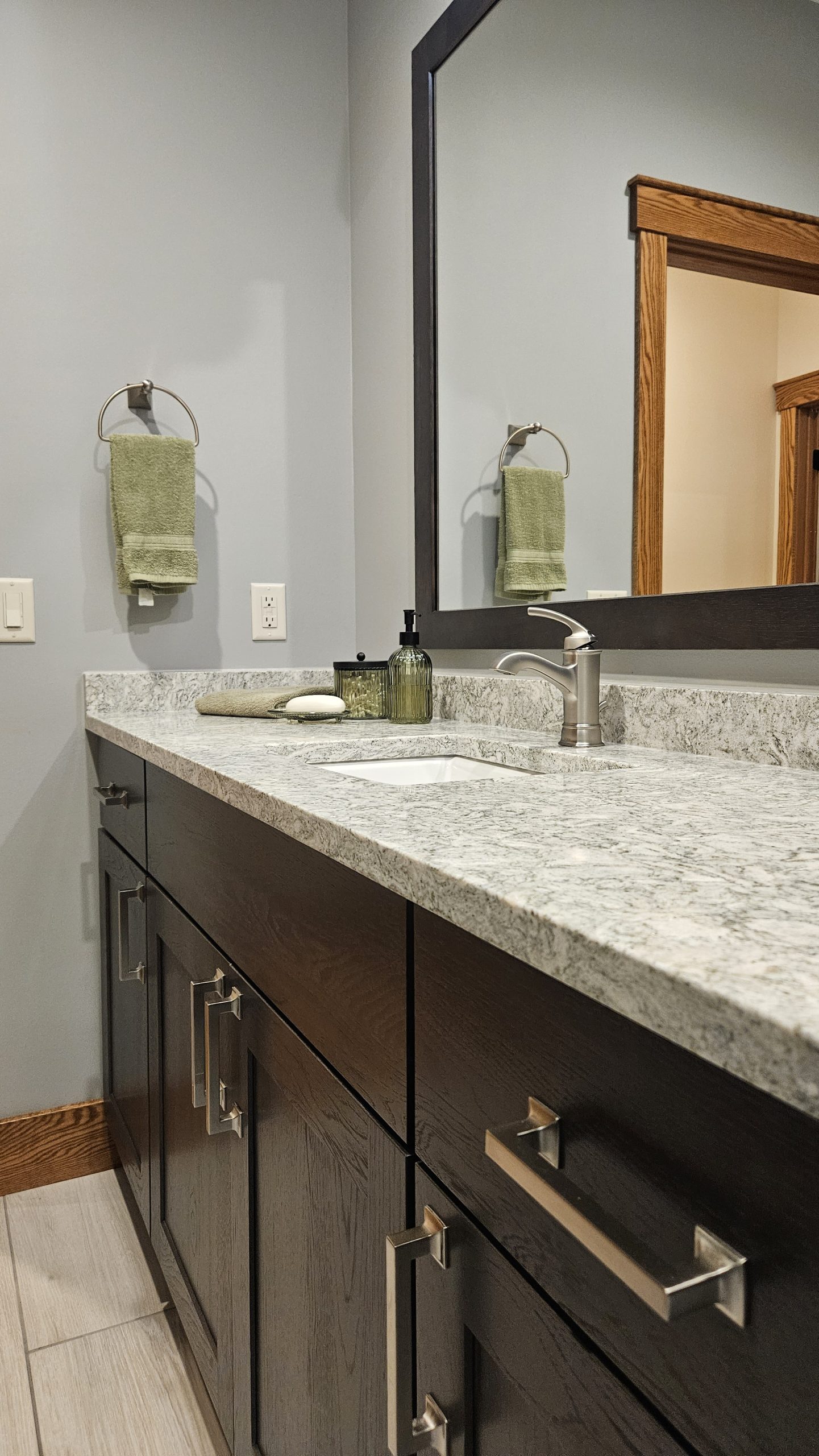 Modern bathroom with a granite countertop, dark wood cabinets, silver fixtures, large mirror, green towel on a ring, soap dispensers near the sink, and custom tile showers for a sleek and personalized touch.