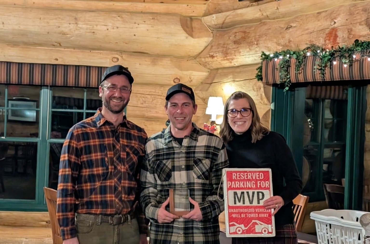 Three people stand indoors; the person in the center holds a trophy, while the person on the right proudly displays a "Reserved Parking for MVP" sign. All are smiling, perhaps celebrating their success.