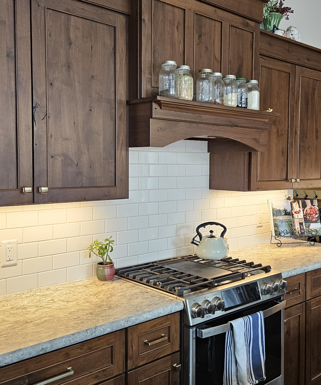 A kitchen with wooden cabinets, white tile backsplash, gas stove with a kettle, jars on a shelf, and potted plant on a light-colored countertop—beautifully designed to complement homes featuring custom tile showers.