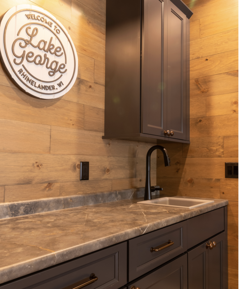A countertop with a sink and dark cabinetry below, set against a wooden wall with a round "Lake George, Rhinelander, WI" sign—perfectly complementing spaces designed for custom tile showers.