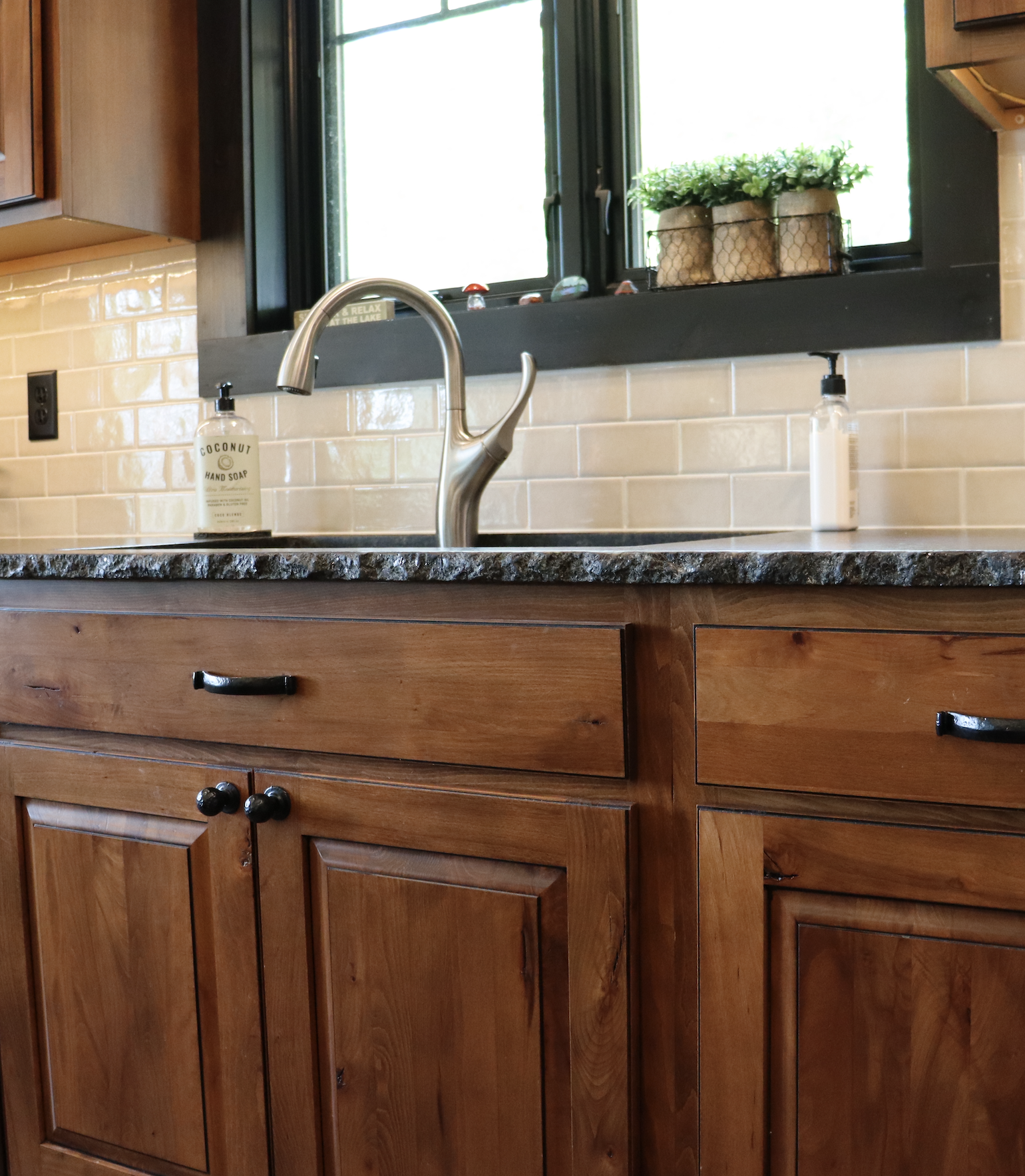 Kitchen sink with a silver faucet, soap dispensers, and wooden cabinets below; light-colored tile backsplash—perfectly complementing custom tile showers—and a window with plants above.