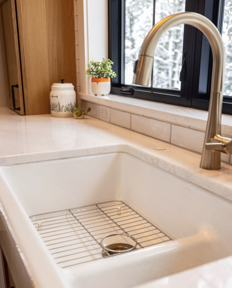 A modern kitchen sink with a stainless steel faucet, white countertop, potted plant, and ceramic jar near a window showing a snowy scene—perfectly complementing homes featuring custom tile showers.