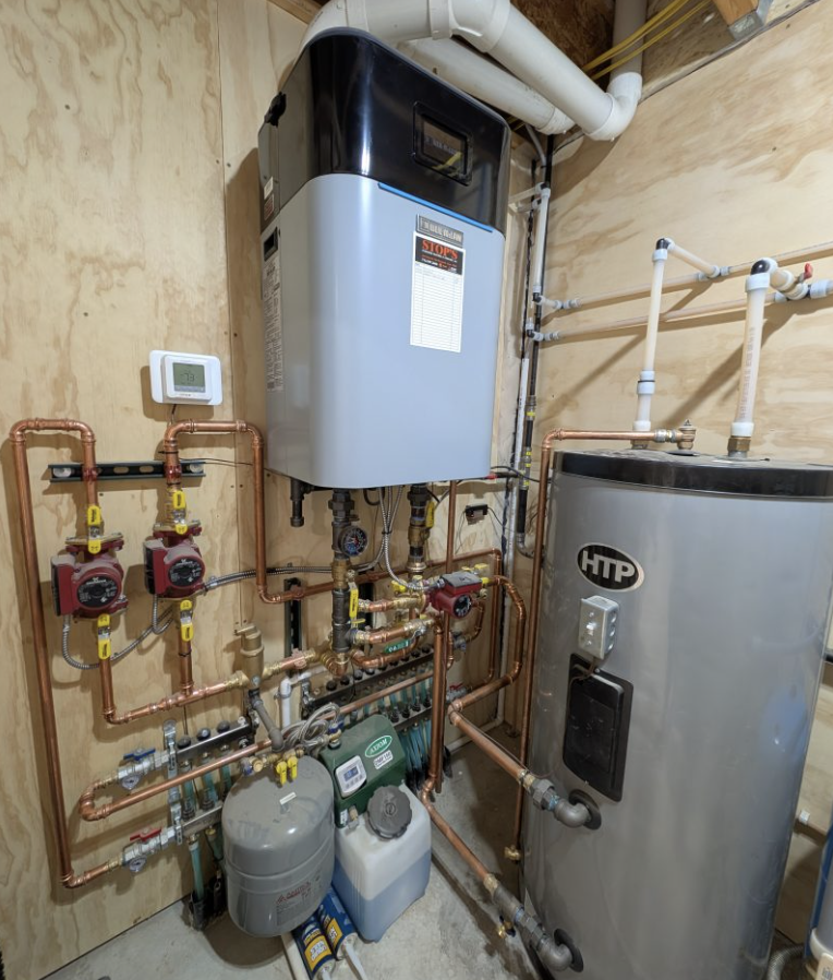 A residential boiler and hot water system with copper piping, pumps, a water tank, control devices, and pressure tanks installed in a utility room with wooden walls—ideal for homes with Custom Tile Showers needing reliable hot water.