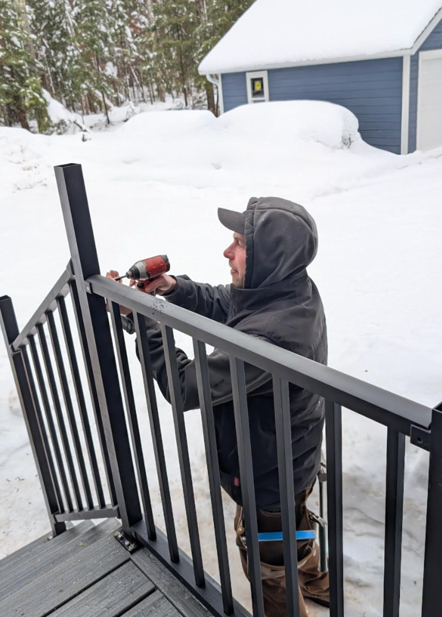 A person in winter clothing uses a power drill to install or repair a metal railing on outdoor steps in a snowy yard near a blue garage.