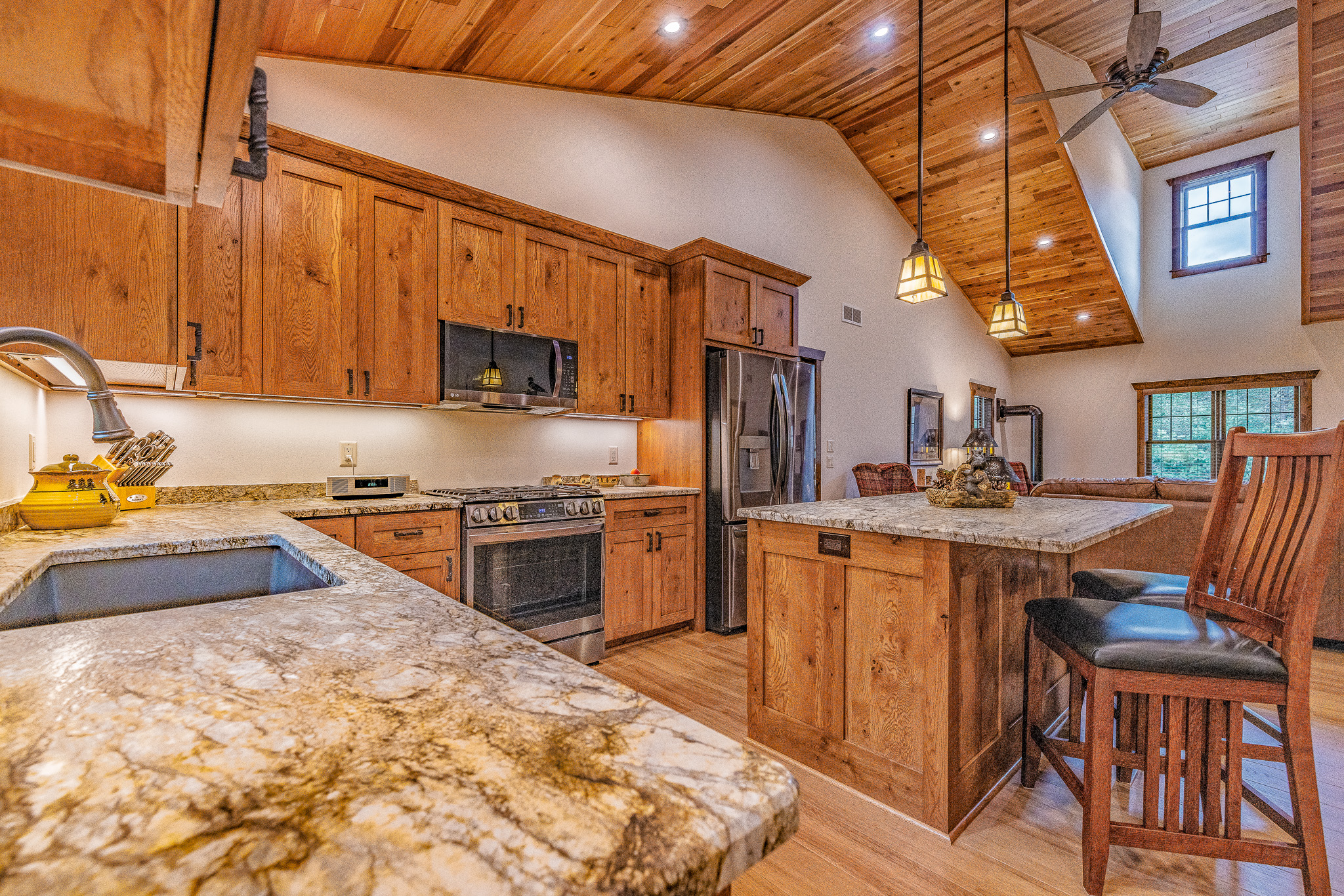 Modern kitchen with wooden cabinets and stainless steel appliances, featuring a marble island and two bar stools under warm ceiling lights in an open-plan living space, complemented by nearby custom tile showers for a touch of luxury.