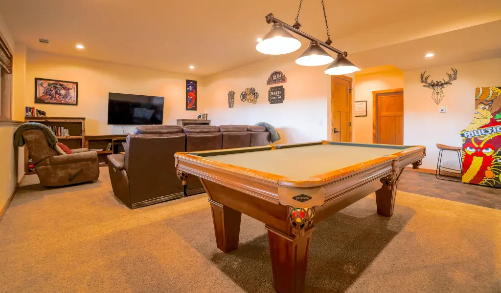 A basement game room transformed by home remodeling, featuring a pool table in the foreground, a seating area with recliners, TV, wall art, and bright overhead lighting.
