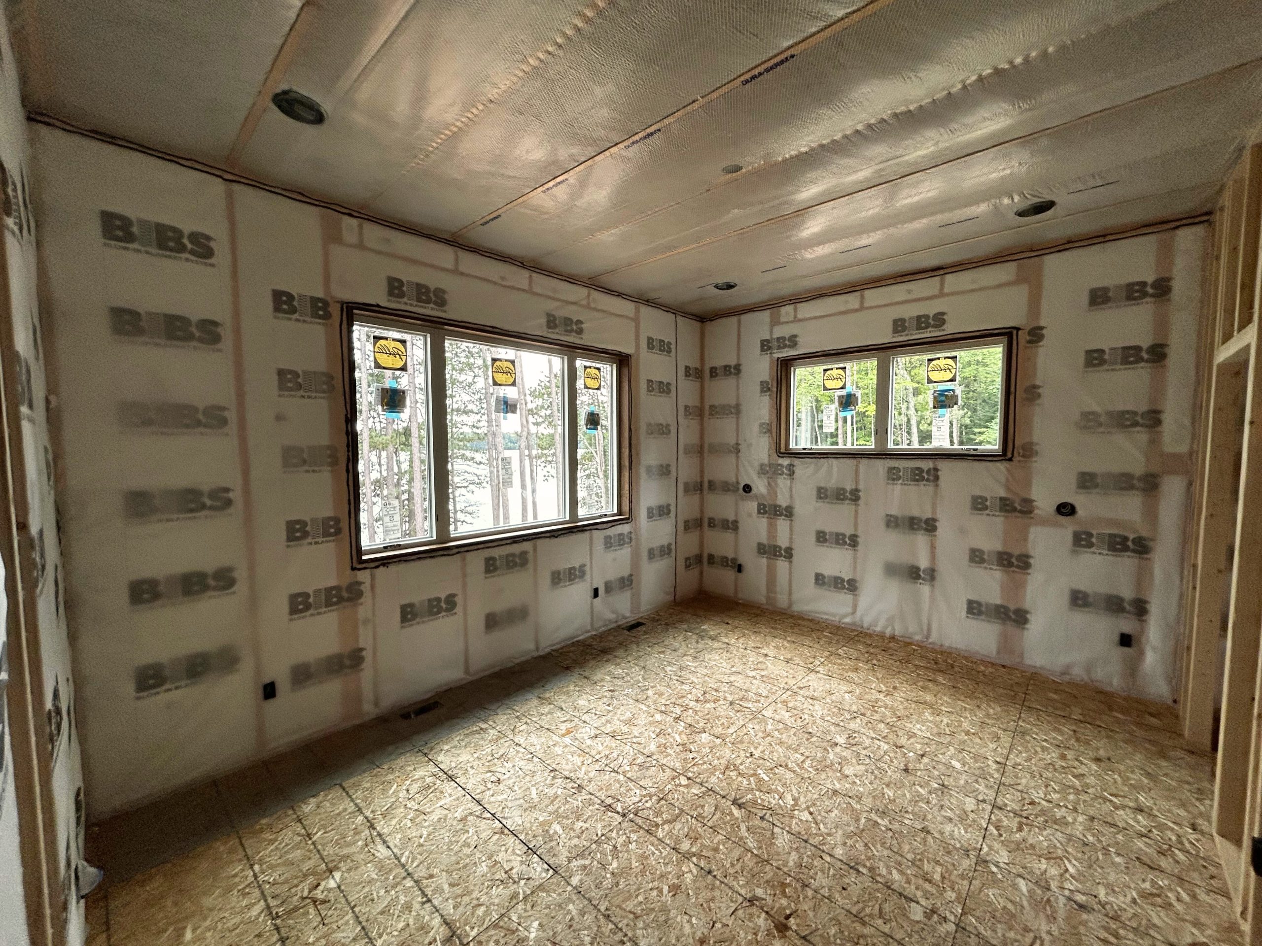 Unfinished room with insulation on walls and ceiling, two large windows, exposed plywood floor—perfect space to design custom tile showers or other personalized features.