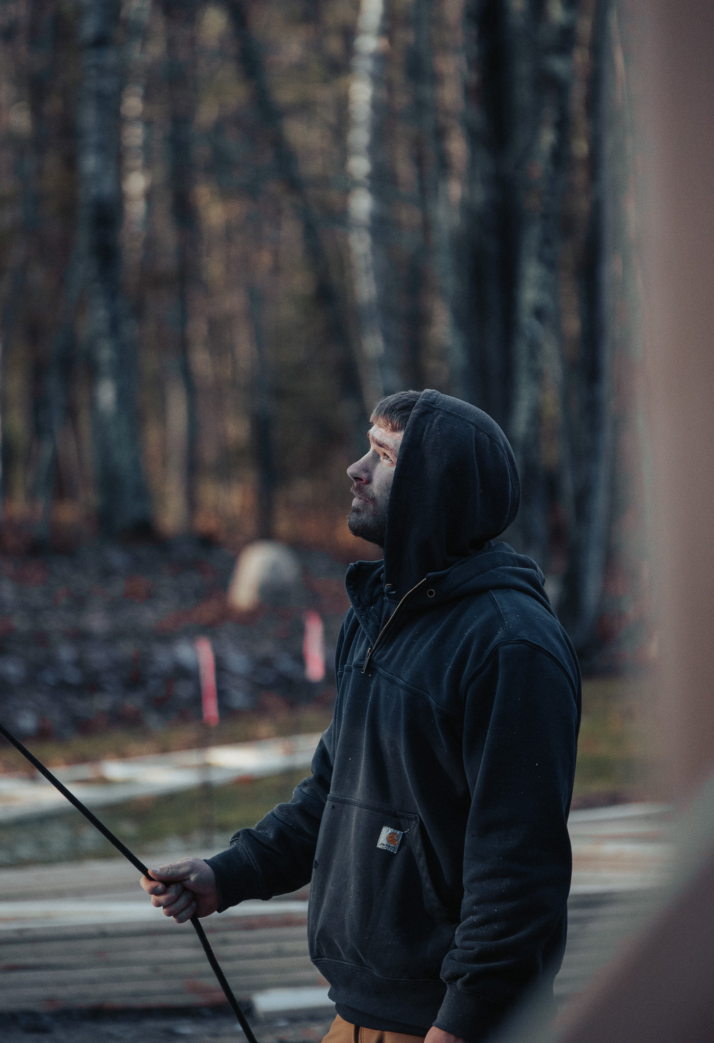 A man in a dark hoodie looks upward while holding a pole or rod outdoors, with trees, construction materials, and hints of custom tile showers in the background.