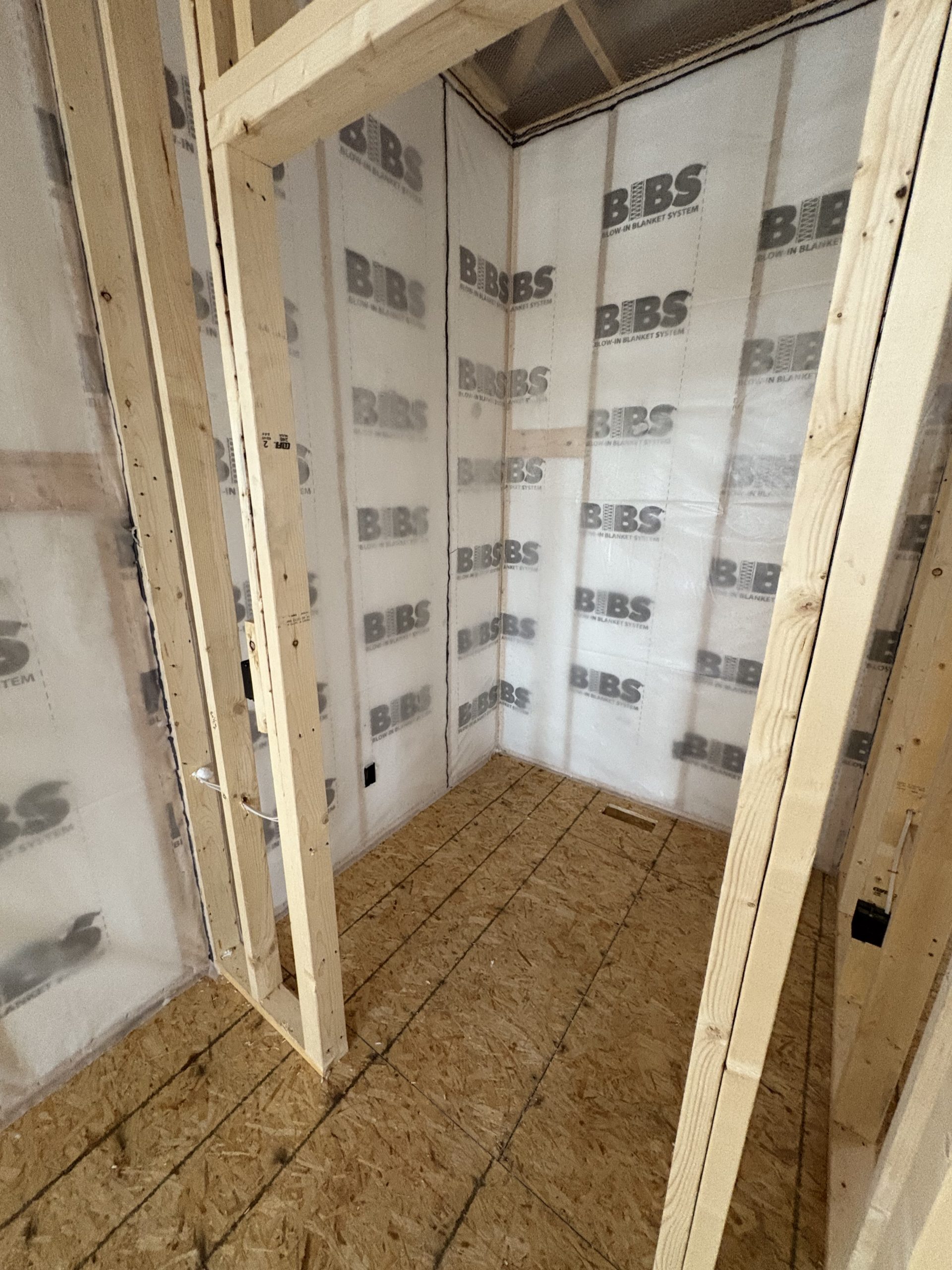 Wood framing of a small room under construction with insulation wrap labeled “BIBS” on the walls and an unfinished plywood floor, ideal for future additions like custom tile showers.