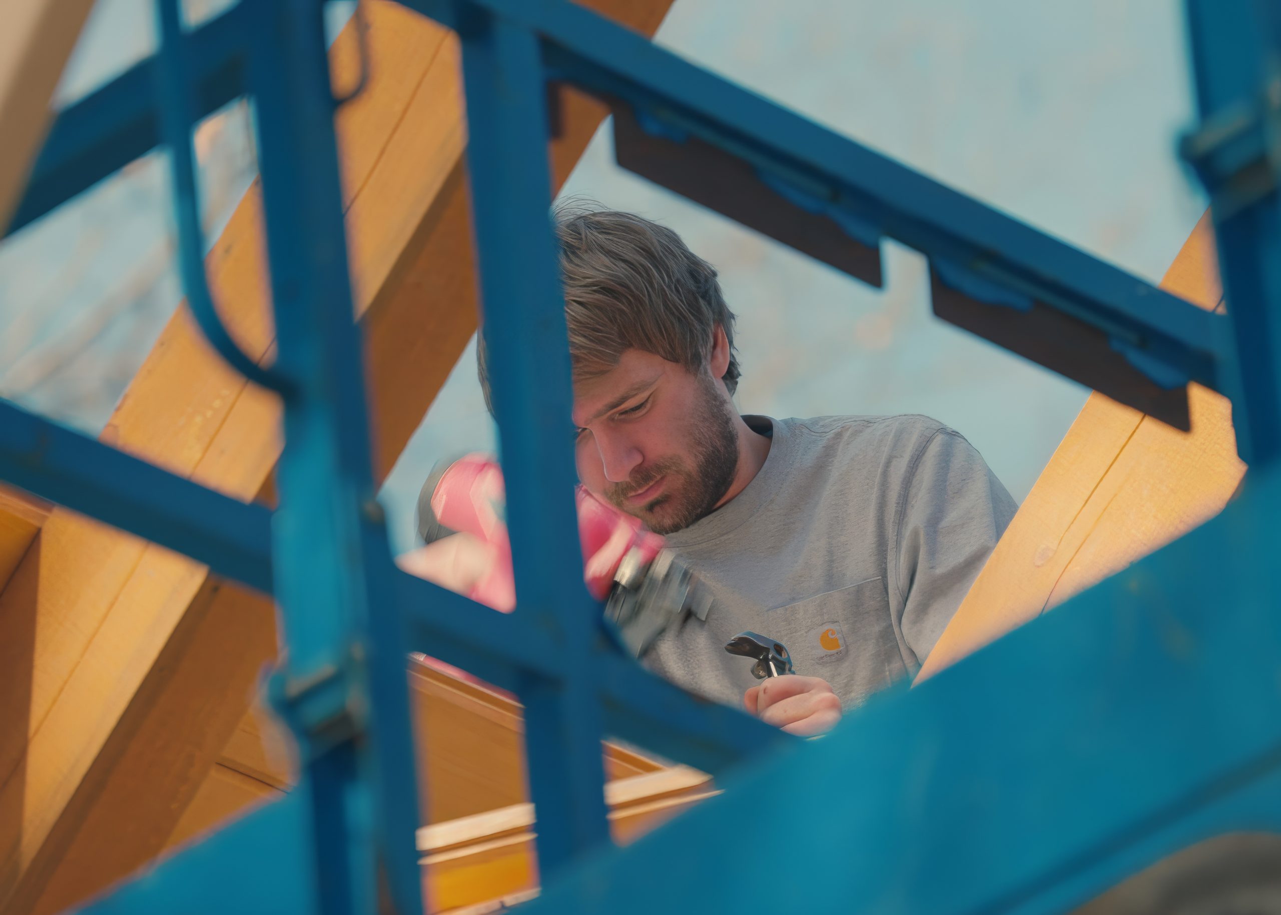 A man using a power drill works on a wooden structure, partially obscured by blue metal framing in the foreground—an early step in crafting custom tile showers.