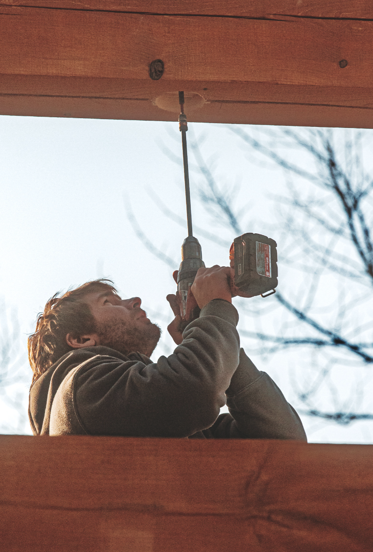 A person uses a cordless drill to drive a screw into a wooden beam overhead, viewed from below with trees visible in the background—perhaps prepping for custom tile showers or other home renovations.