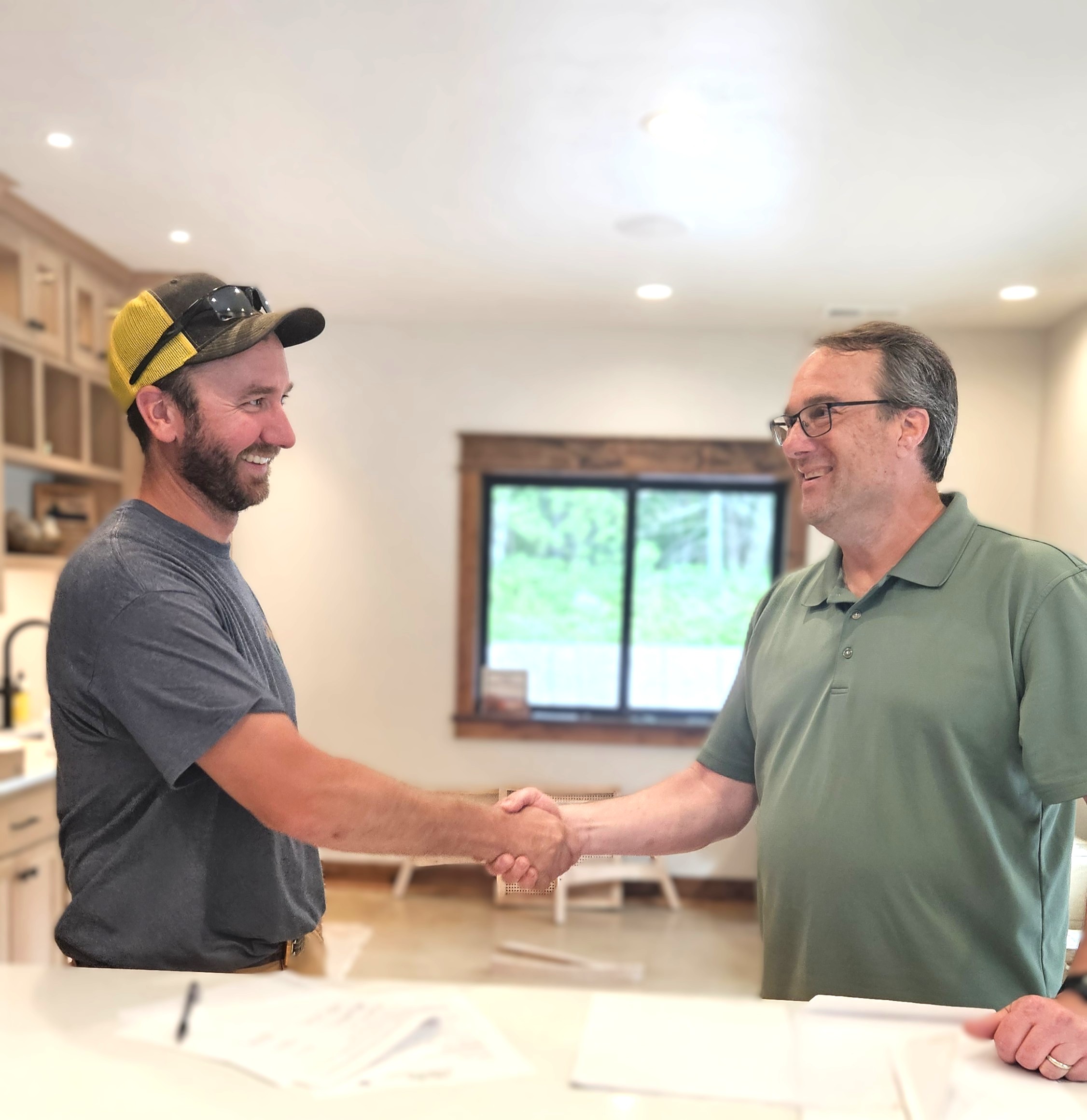 Two men shake hands and smile at each other in a bright kitchen, with paperwork on the counter in front of them, discussing their new custom tile showers.