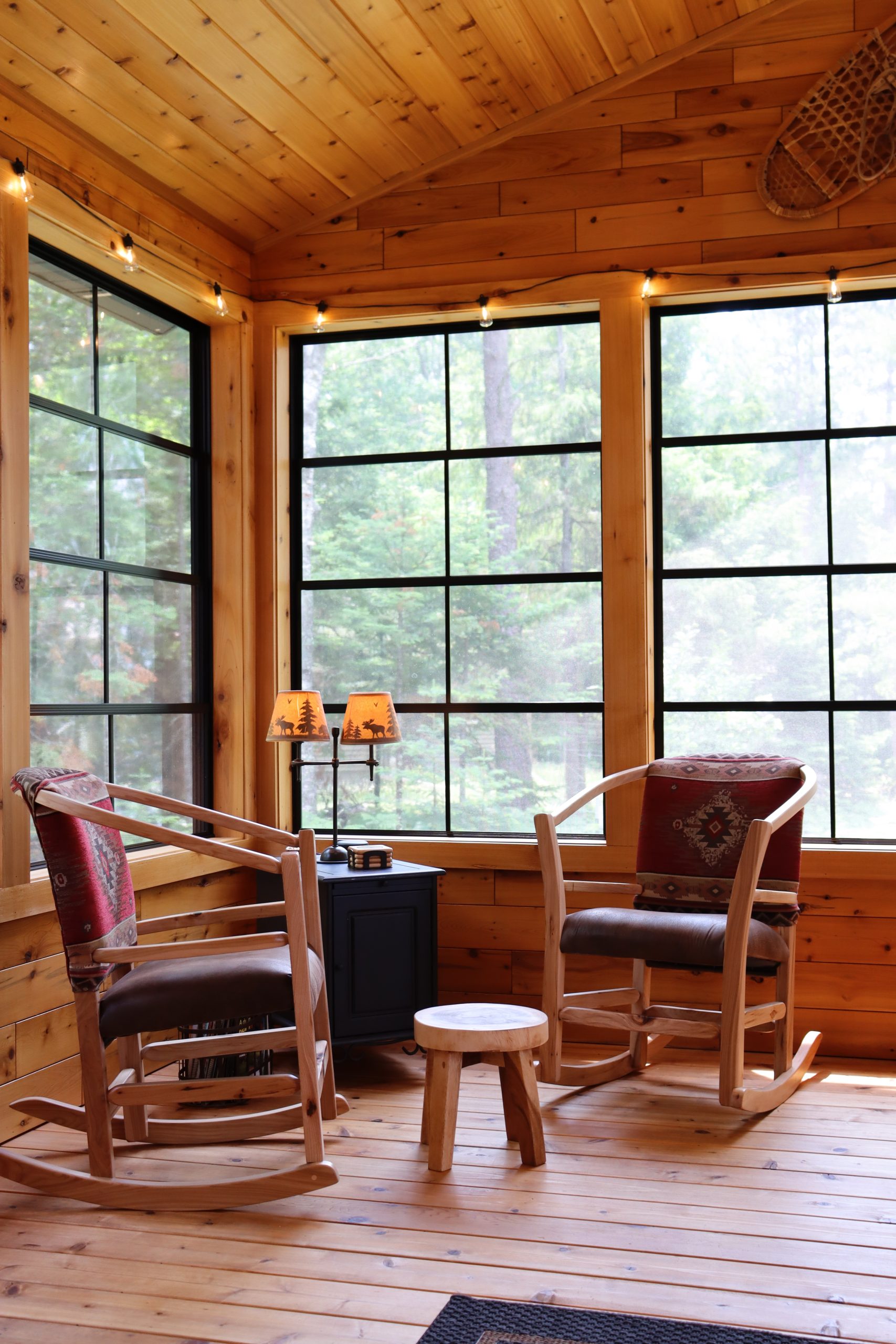 A cozy wooden sunroom with two cushioned rocking chairs, a small table with two lamps, large windows overlooking trees outside, and easy access to custom tile showers for added comfort.