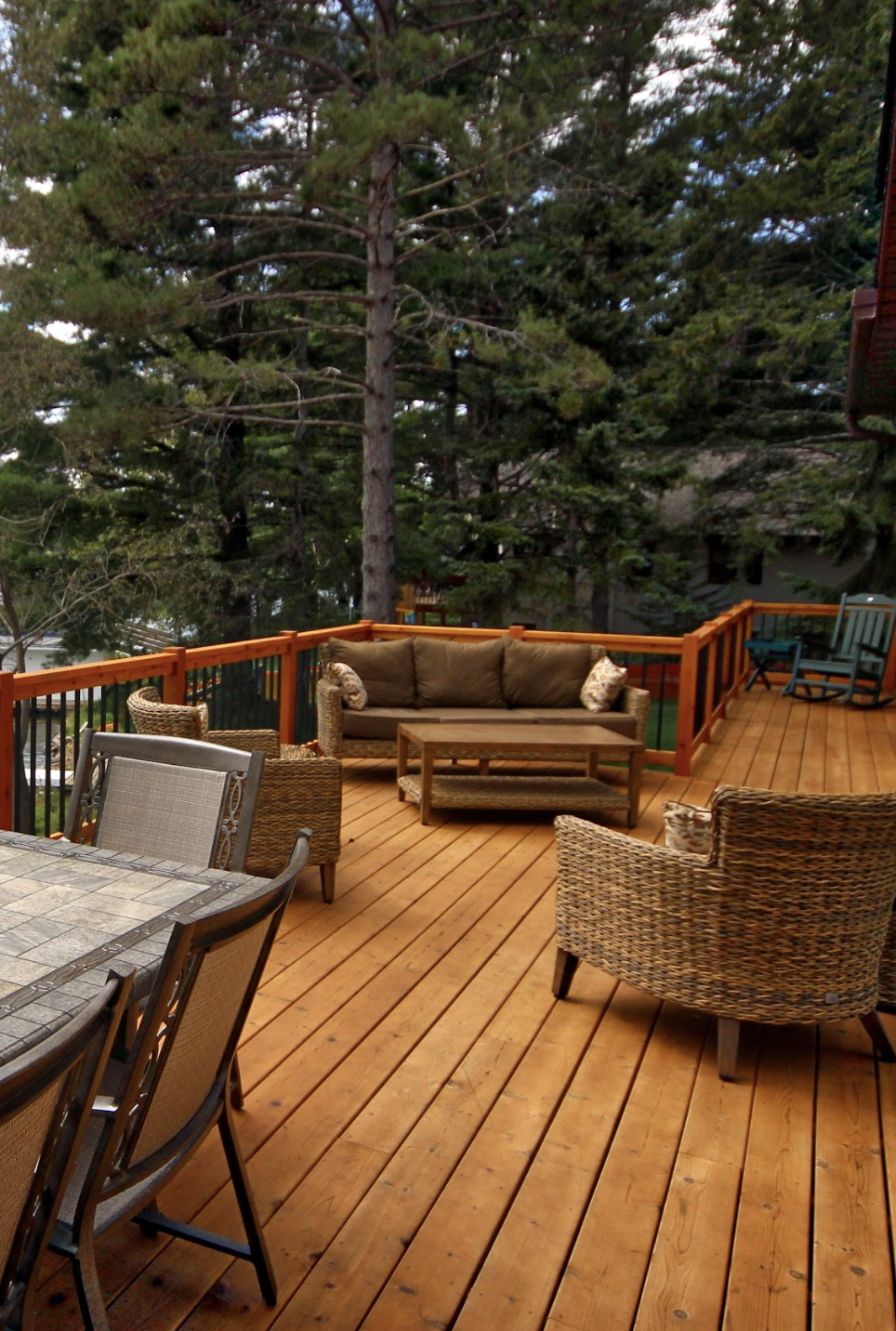 A wooden deck with wicker and cushioned seating, a dining table, and chairs, surrounded by a wooden railing and tall pine trees—ideal for relaxing after enjoying custom tile showers indoors.