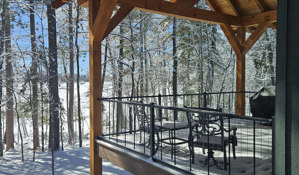 A snow-covered patio with metal table and chairs overlooks a forest of tall trees, all blanketed in fresh snow under a clear blue sky—perfect inspiration for designing custom tile showers with serene, natural themes.