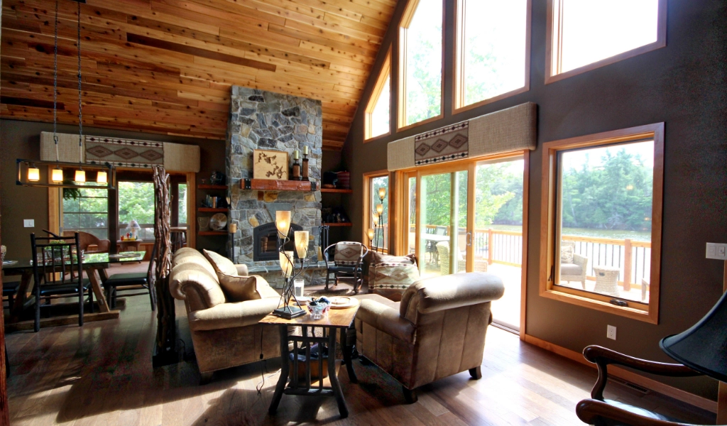 A spacious living room with large windows, wood ceiling, stone fireplace, brown sofas, and natural light overlooking a deck and trees outside—plus custom tile showers for a touch of luxury throughout the home.