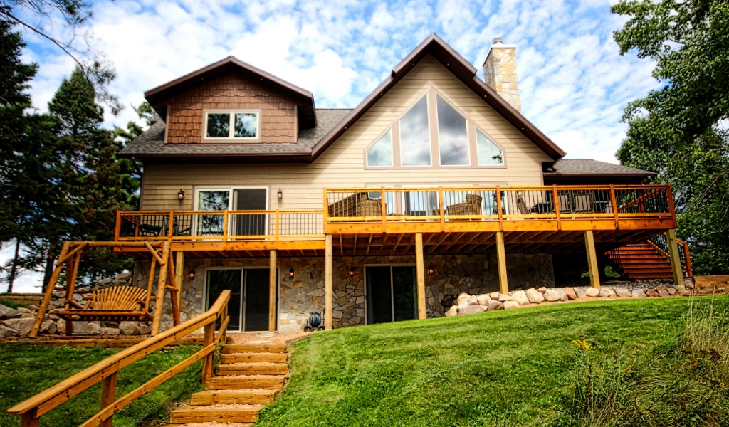 A two-story house with large triangular windows, wooden deck, stone foundation, and a swing sits on a grassy lawn with trees in the background—featuring custom tile showers for a touch of luxury inside.